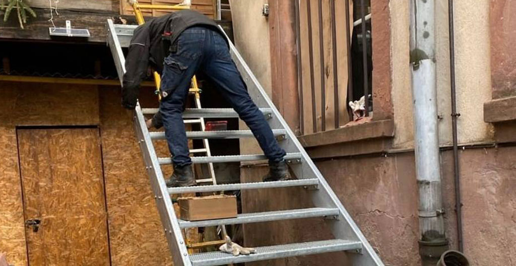 Deux techniciens professionnels installant escalier extérieur aluminium préfabriqué sur façade duplex montréalais, un stabilisant structure depuis haut, l'autre fixant ancrages muraux avec perceuse, contexte chantier actif